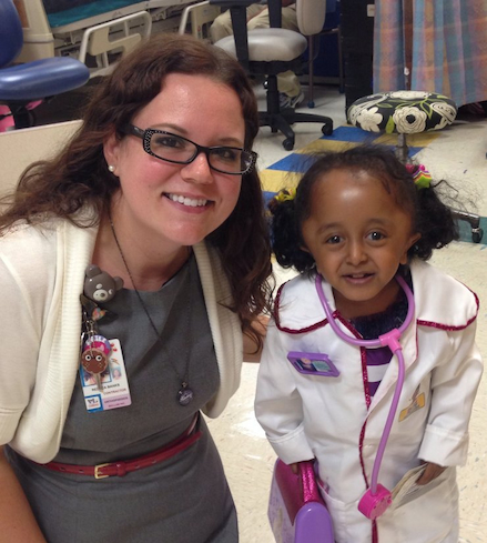 Rebeca smiling with her patient who is dressed as Doc McStuffins in a white labcoat with pink stethoscope